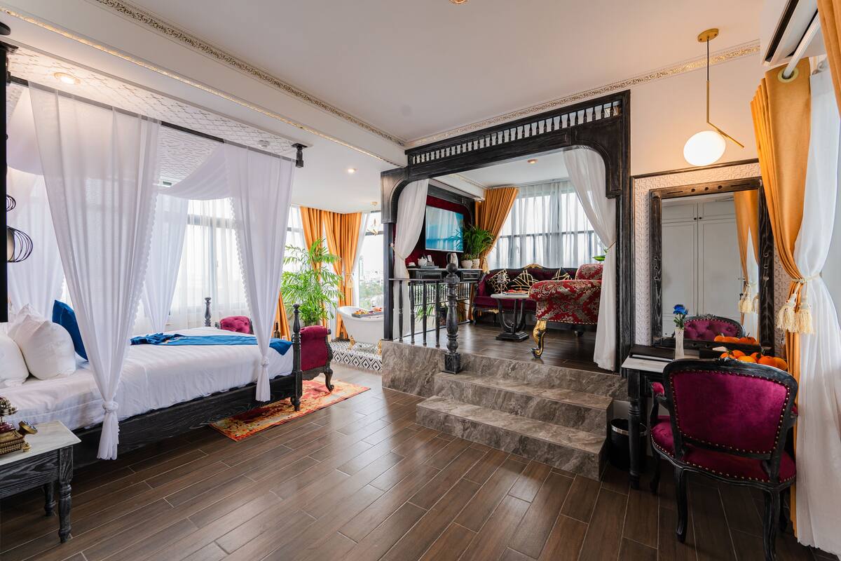 Um quarto luxuoso com uma cama de dossel, um espaço de estar com sofá e poltronas, um espelho grande e vista para a cidade.