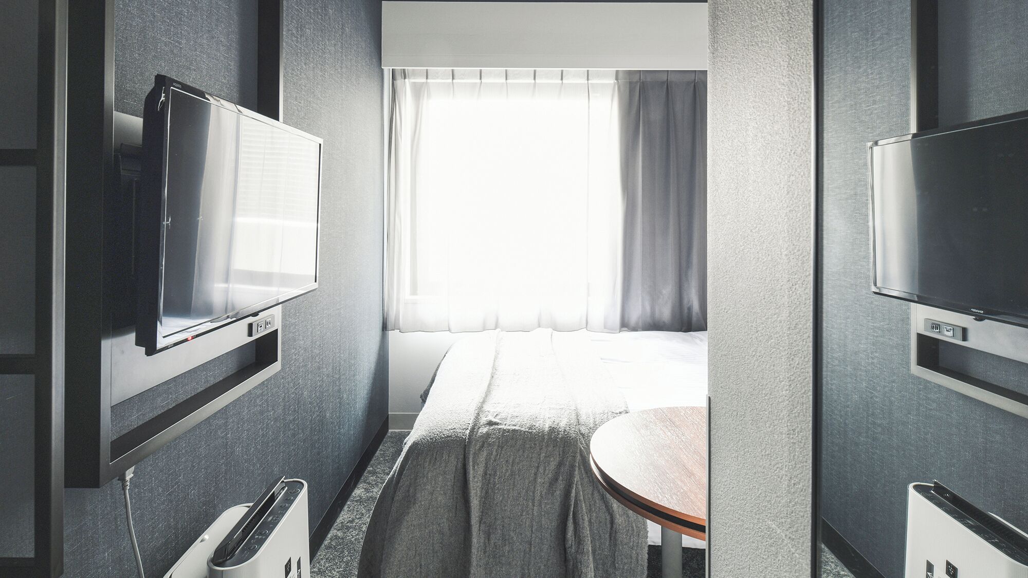 Una habitación de hotel con cama, televisor montado en la pared, ventana con cortinas y una mesita.