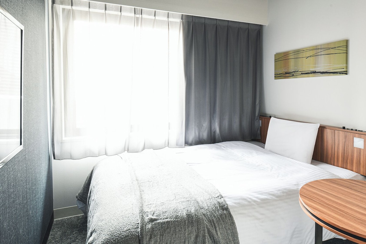 Habitación de hotel con cama, cabecero de madera, una mesita y una ventana con cortinas.