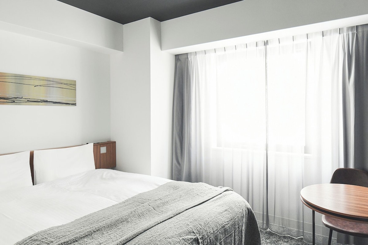 Habitación de hotel con cama, una mesita, una silla, ventana con cortinas y una obra de arte en la pared.
