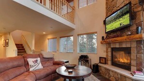 A living room with a stone fireplace, a television mounted above it, a brown leather sofa, and a staircase leading to an upper level. - Bear Mtn Chalet |Walk 2 Zoo, Bear Mtn & Golf+Games (Big Bear Lake)