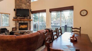 A living room with a stone fireplace, a flat-screen TV, a dining table, and large windows. - Bear Mtn Chalet |Walk 2 Zoo, Bear Mtn & Golf+Games (Big Bear Lake)