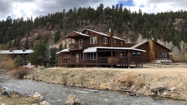 A two-story wooden cabin with a deck, a river in the foreground, and a forested hillside in the background. - KXDJ Riverside 1 on the River (Red River)