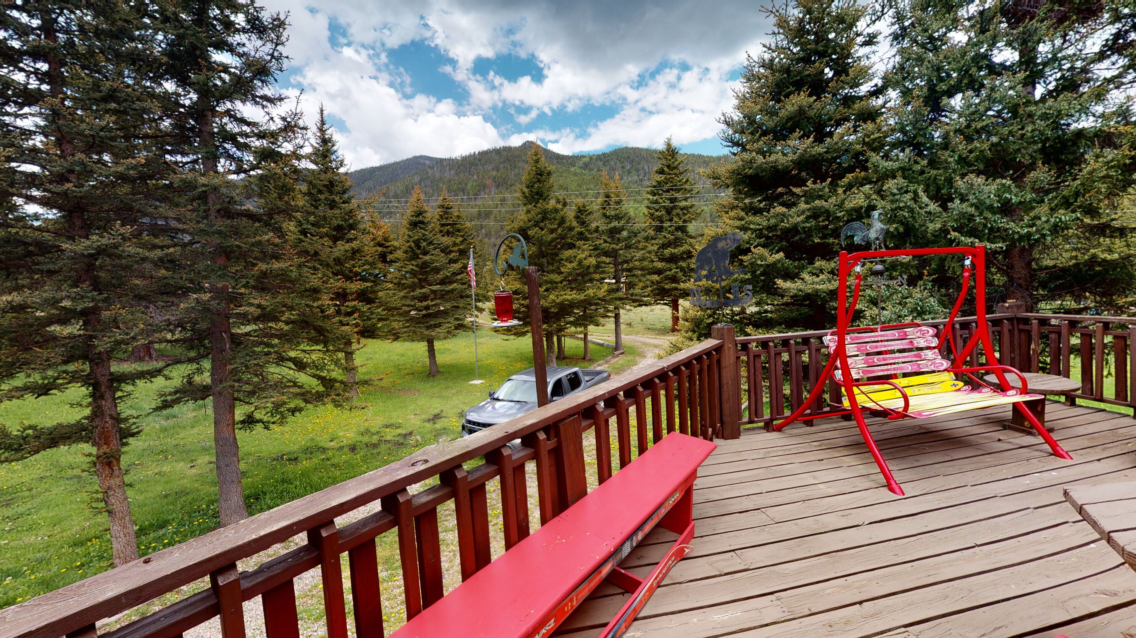 Une terrasse en bois avec un banc rouge et une balançoire, donnant sur une zone herbeuse avec des arbres et une montagne en arrière-plan.