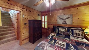 Interior de una cabaña con escalera de madera, cama con colcha, cómoda y ventilador de techo.