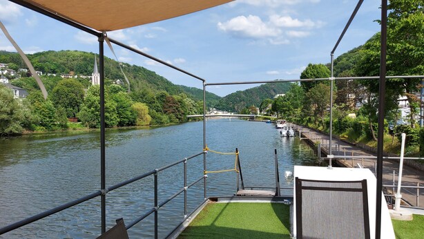 Blick auf einen Fluss von einer überdachten Terrasse mit Geländer, einer Rasenfläche und einer Bank.
