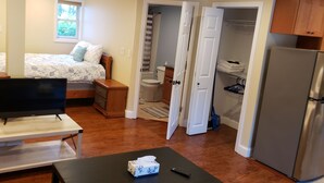 Ein kompakter Wohnbereich mit einem Bett, einem Fernseher auf einem Lowboard, einem kleinen Tisch mit Taschentuchbox und Fernbedienung, einer Küchenzeile mit Kühlschrank und einem Badezimmer mit Duschvorhang.