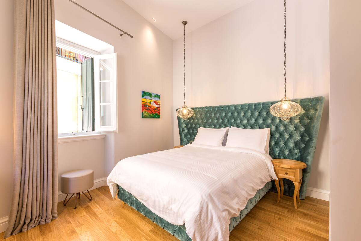 Un dormitorio con una cama grande, una ventana con cortinas, una mesita de noche pequeña y una lámpara colgante.