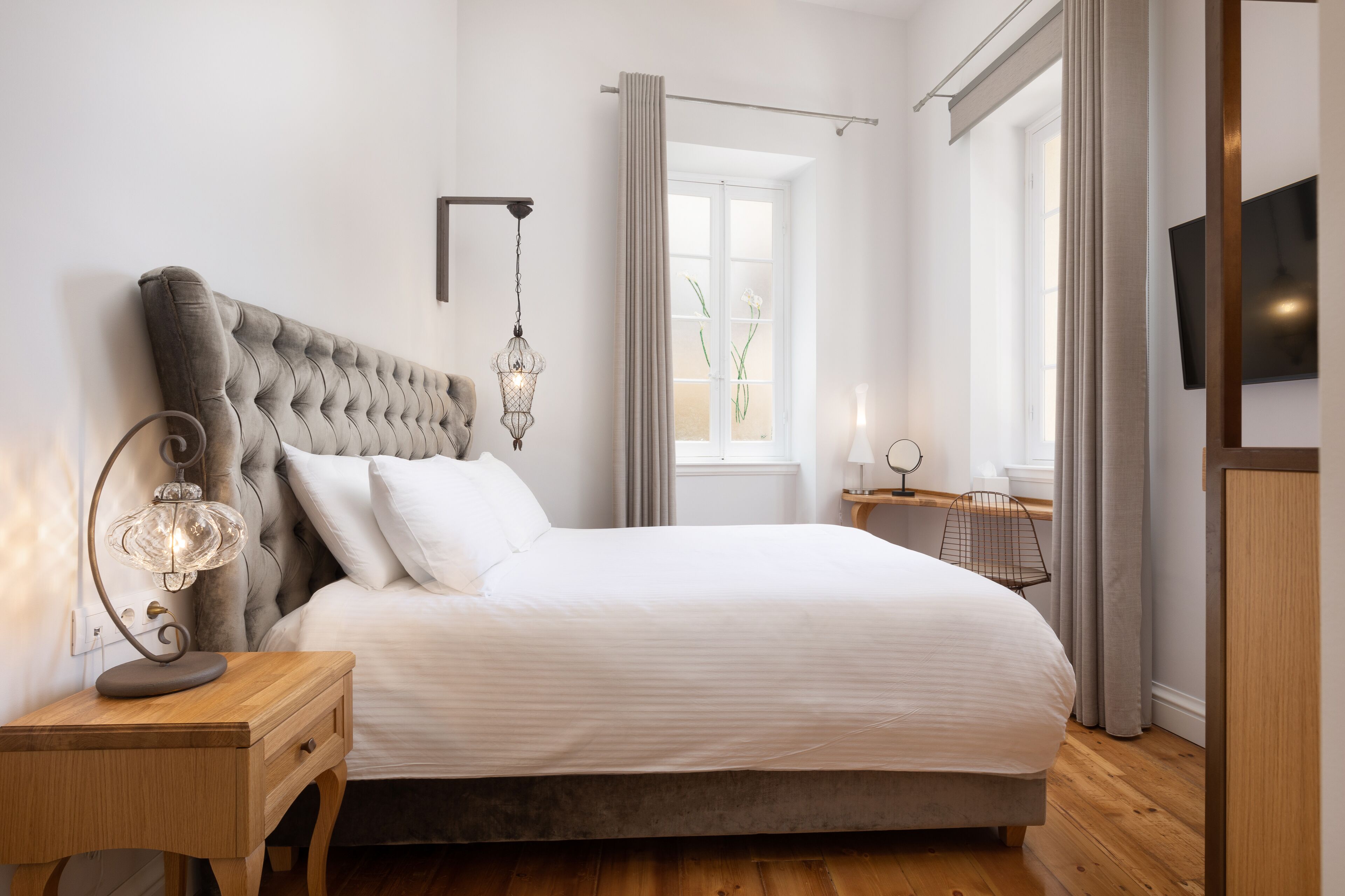 Une chambre à coucher bien aménagée, avec une tête de lit capitonnée, une table de chevet avec une lampe, une fenêtre avec des rideaux et un téléviseur à écran plat fixé au mur.