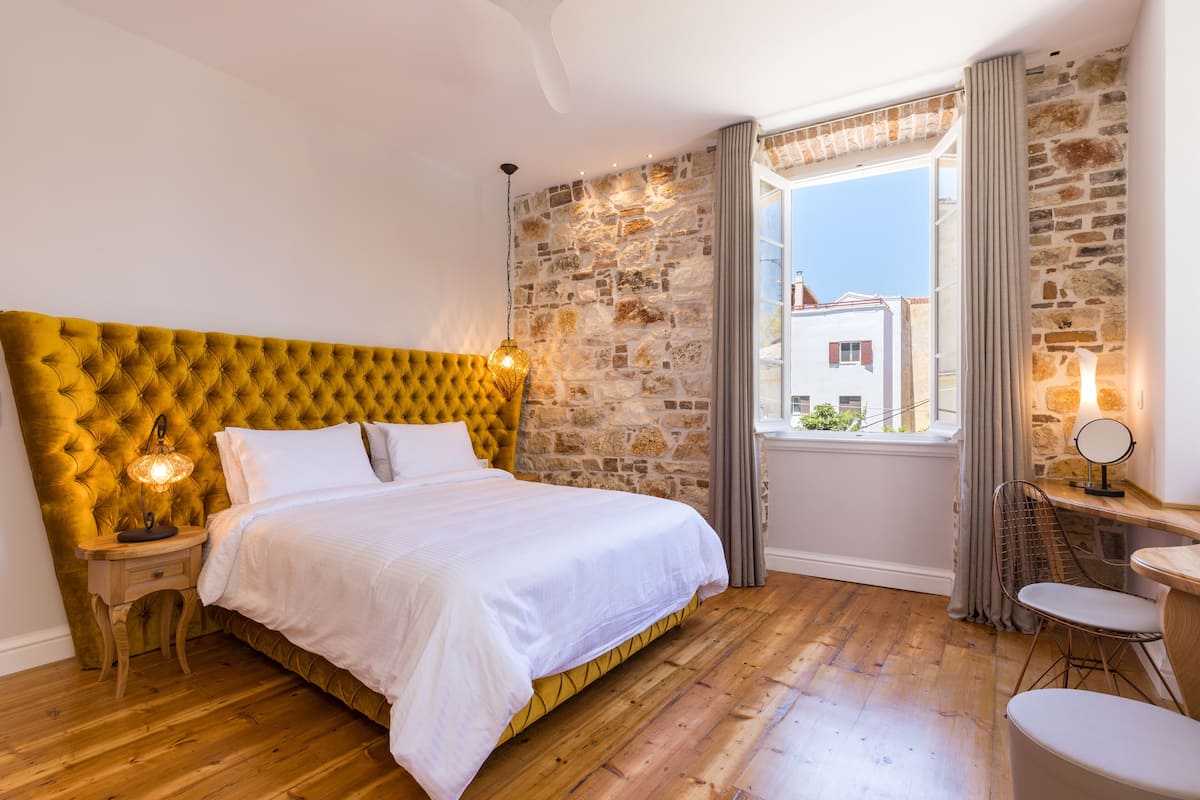 Un dormitorio con cabecera tapizada de color amarillo, suelo de madera, cama blanca, mesita de noche con lámpara, ventana con cortinas y una pequeña mesa con una silla.
