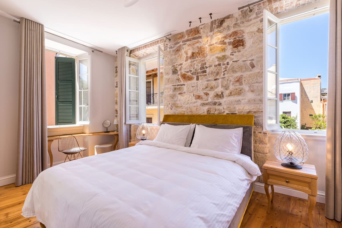 Un dormitorio con pared de piedra, una cama de madera, una mesita de noche con lámpara y una ventana con cortinas.