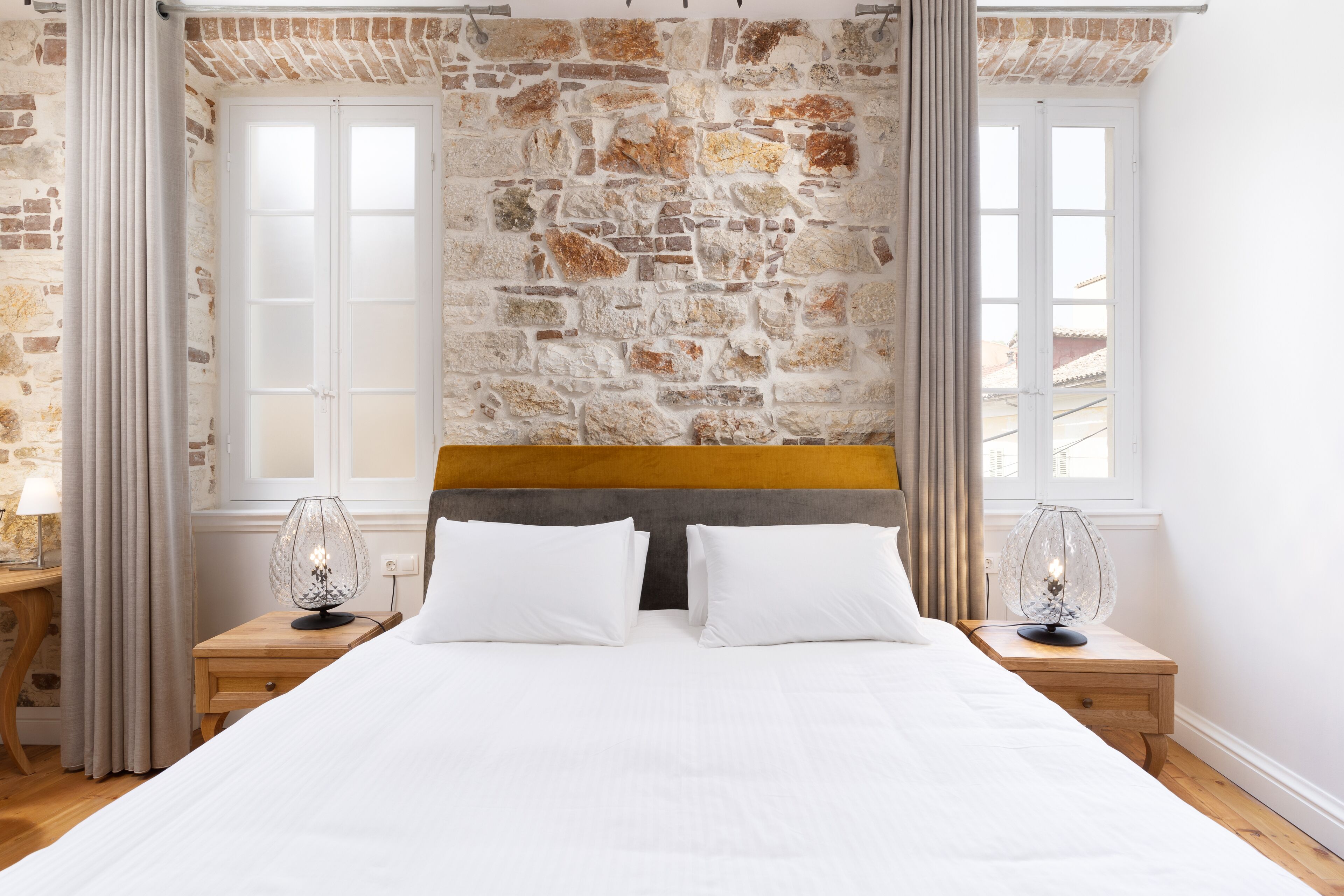 Een slaapkamer met een stenen muur, een houten hoofdbord, een bed met witte beddengoed, twee bedlampen en een raam met gordijnen.