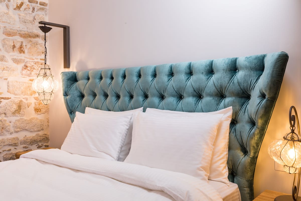 Una cama con cabecera capitonada, ropa de cama blanca y una pared de piedra.