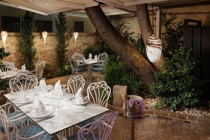 Un área de comedor al aire libre con sillas y mesas de metal blanco dispuestas para una comida, rodeada de follaje y un muro de ladrillo.