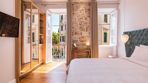 A bedroom with a large bed, a stone wall, a wooden floor, and a balcony with a view of buildings. - Acanthus blue (Corfu)