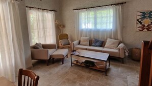 Un salón acogedor con un sofá, sillones y una mesita de centro. Hay dos ventanas con cortinas, techo de madera y suelo de baldosas.
