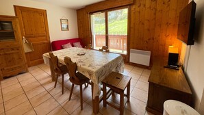 A cozy room with a wooden interior, a dining table with chairs, a red sofa, a television, and a window with a view of greenery. - Cozy 2-bed apt at ski slopes with parking and WiFi (Flumet)