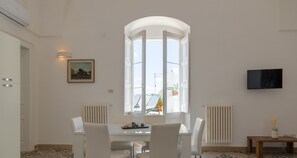 Un espace repas moderne avec une table et des chaises blanches, une grande fenêtre offrant une belle vue et un téléviseur fixé au mur.