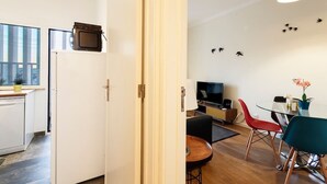 Een moderne keuken met een inbouwvaatwasser, een eethoek met een ronde tafel en rode stoelen, en een woonruimte met een televisie en een wandlamp.
