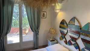 Una habitación con cama, ventana con cortinas, lámpara y un cuadro llamativo en la pared.