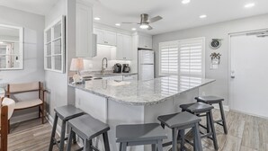 A kitchen with a central island, bar stools, a ceiling fan, and a refrigerator. - ✨Direct Ocean - Chateau by the Sea 13C✨ (New Smyrna Beach)