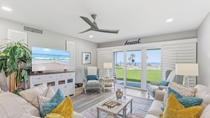 A living room with a television, a ceiling fan, a large window with a view of the beach, and a door leading outside. - ✨Direct Ocean - Chateau by the Sea 13C✨ (New Smyrna Beach)