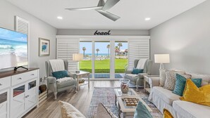 A living room with a ceiling fan, a TV, a sofa, a coffee table, and a view of the beach through the windows. - ✨Direct Ocean - Chateau by the Sea 13C✨ (New Smyrna Beach)