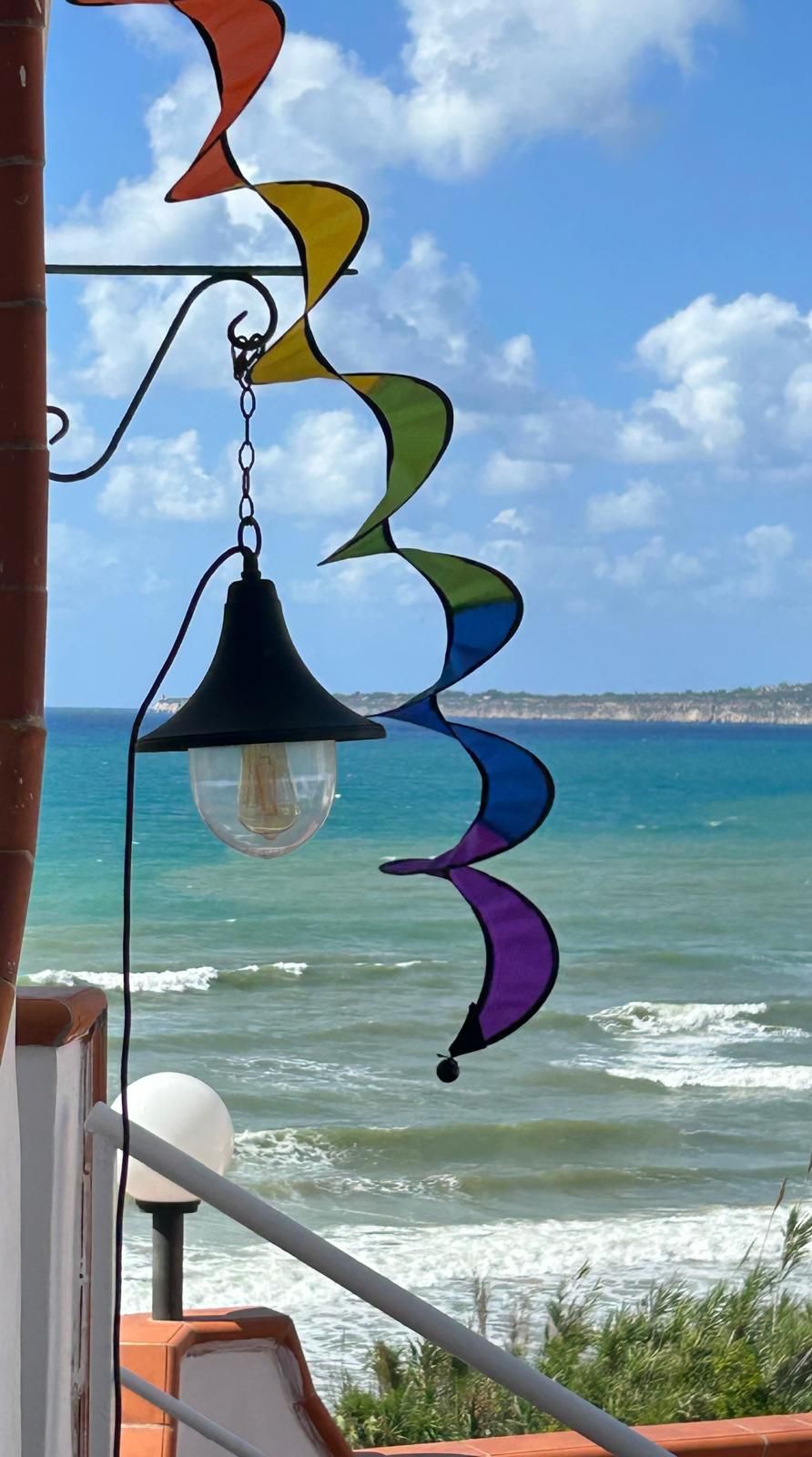 Ein bunter Windspiel hängt an einem Fenster mit Blick auf einen Strand und Wellen.