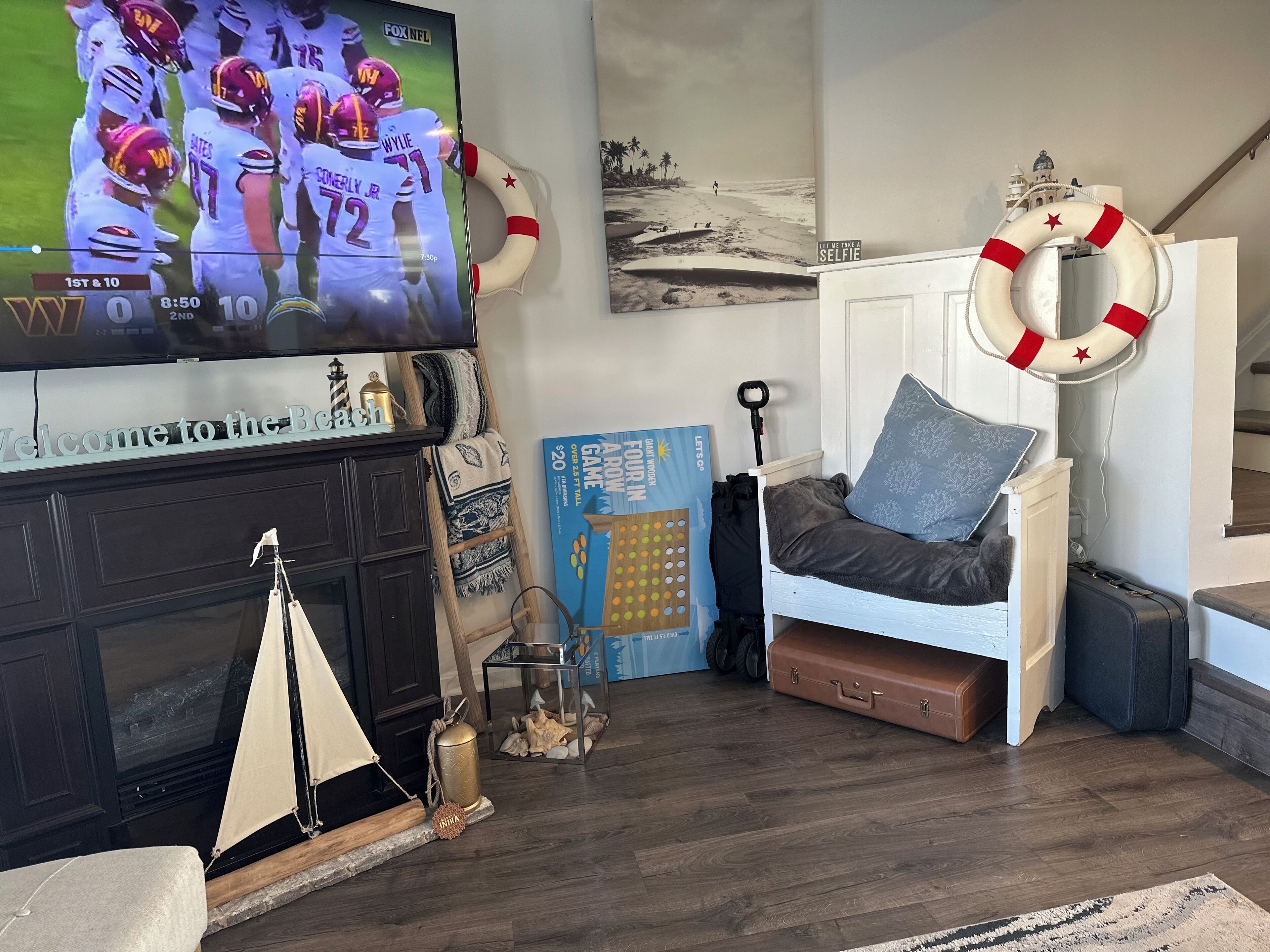 A cozy living room with a TV displaying a football game, a fireplace, a beach-themed painting, and a small ladder.