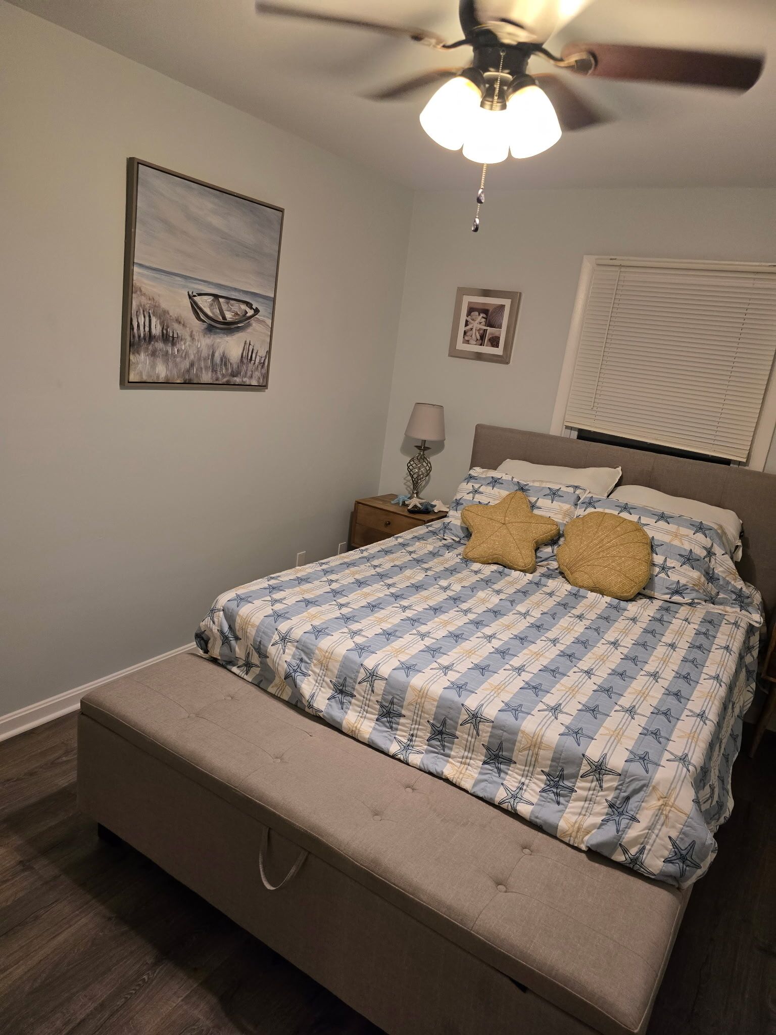 Een slaapkamer met een bed, nachtkastje, plafondventilator, raam met jaloezieën en een schilderij aan de muur.