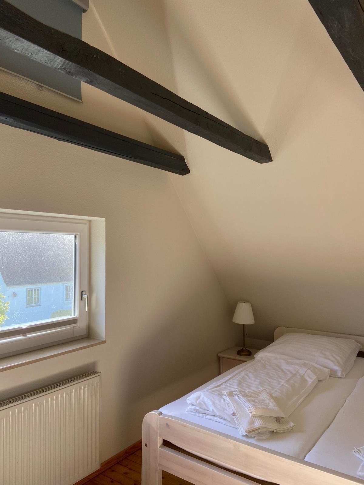 Een slaapkamer met een raam, een radiator, een nachtkastje met een lamp, en een bed met witte lakens.