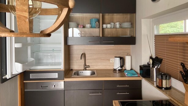 Eine kleine Küche mit Einbauofen, Spüle und Herd. Es gibt Schränke mit Geschirr und Besteck, sowie einen Deckenventilator aus Holz.