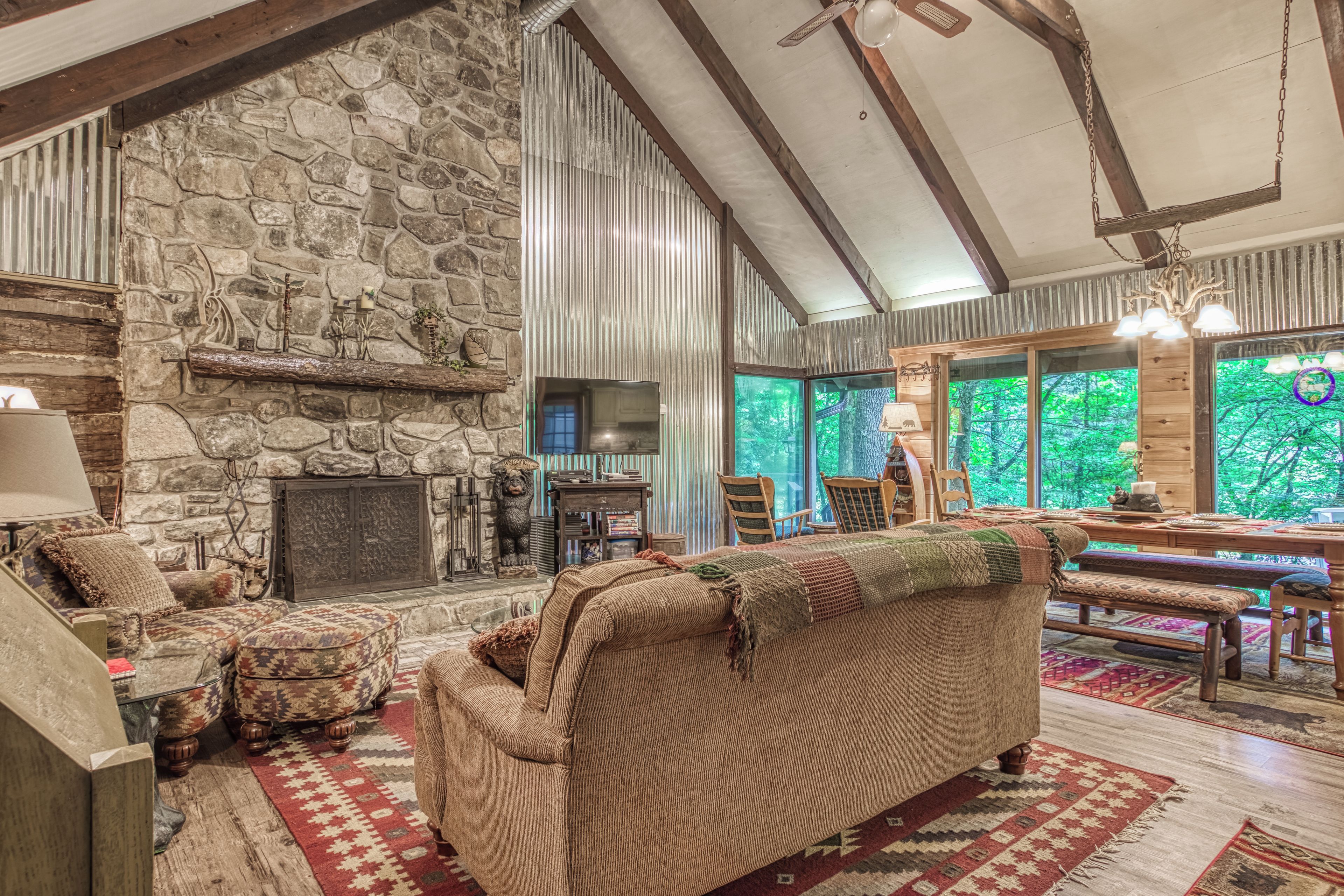 A cozy living room with a stone fireplace, wooden beams, and a large window overlooking greenery.