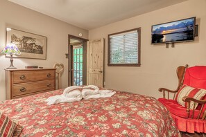 A bedroom with a floral bedspread, a wooden dresser, a red armchair, a television on the wall, and a framed picture. - Creekside | Hot Tub | Wood Burning Fireplace | Fire Pit | Wifi (Gatlinburg)