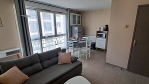 A living room with a sofa, a dining table with chairs, a kitchen area with appliances, and large windows. - One-Bedroom Apartment, good standard,  with balcony and parking (Lyon)