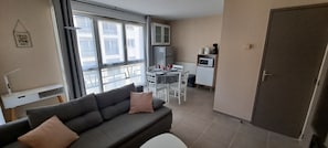 A living room with a sofa, a dining table with chairs, a kitchen area with appliances, and large windows.