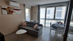 A living room with a grey sofa, a small white table, and a dining area with a table and chairs. - One-Bedroom Apartment, good standard,  with balcony and parking (Lyon)