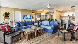 Ein Wohnzimmer mit blauen Sofas, einem Couchtisch und Korbstühlen. Im Hintergrund ist eine Küche mit weißen Schränken und einer Mikrowelle zu sehen.