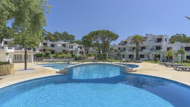 Une piscine au design incurvé, entourée d’un bâtiment blanc doté de balcons et bordée d’arbres.