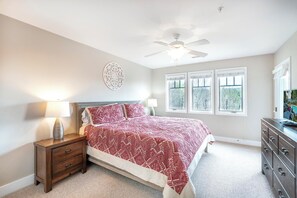 Ein Schlafzimmer mit Bett, Nachttischen, Kommode, Deckenventilator und Fenstern.