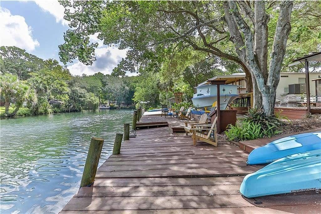 Un muelle de madera que se adentra en un río tranquilo, con kayaks y una zona de estar cubierta bajo los árboles.