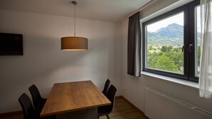 A dining area with a wooden table and chairs, a hanging light fixture, a window with curtains, and a view of greenery and mountains. - 4805 Cirrus 5 (St. Gallenkirch)