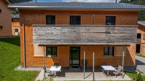 Un bâtiment moderne à deux étages, doté d’une façade en bois, d’un espace extérieur couvert avec des tables et des chaises, et offrant une vue sur un paysage montagneux.