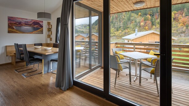 A modern interior with a dining area, a large sliding glass door leading to an outdoor deck with a view of cabins and autumn foliage. - 46142 Montan 14 OG (St. Gallenkirch)