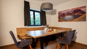 En matplats med ett träbord, fyra grå stoppade stolar, en hängande lampa, ett fönster med mörka gardiner och en tavla som föreställer ett bergigt landskap på väggen.