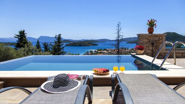 A poolside view with a clear view of the sea and mountains, a table with a hat, a fruit bowl, and two glasses of orange juice. - Villa Kallisto with Infinity Pool & Stunning Sea View in Perigiali, Lefkada!!! (Perigiali)