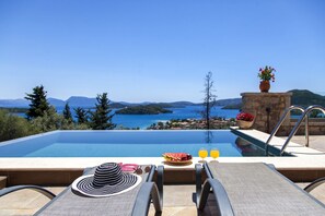 A poolside view with a clear view of the sea and mountains, a table with a hat, a fruit bowl, and two glasses of orange juice.