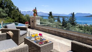 A terrace with wicker furniture, a table with breakfast items, and a view of a lake and mountains. - Villa Kallisto with Infinity Pool & Stunning Sea View in Perigiali, Lefkada!!! (Perigiali)