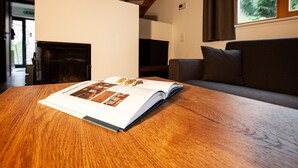 Een open boek op een houten tafel in een kamer met een bank, een open haard en een raam met uitzicht op bomen.