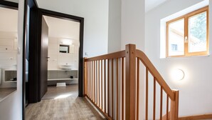 Een hal met houten leuning, een badkamer met wastafel en spiegel, en een raam met natuurlijk licht.